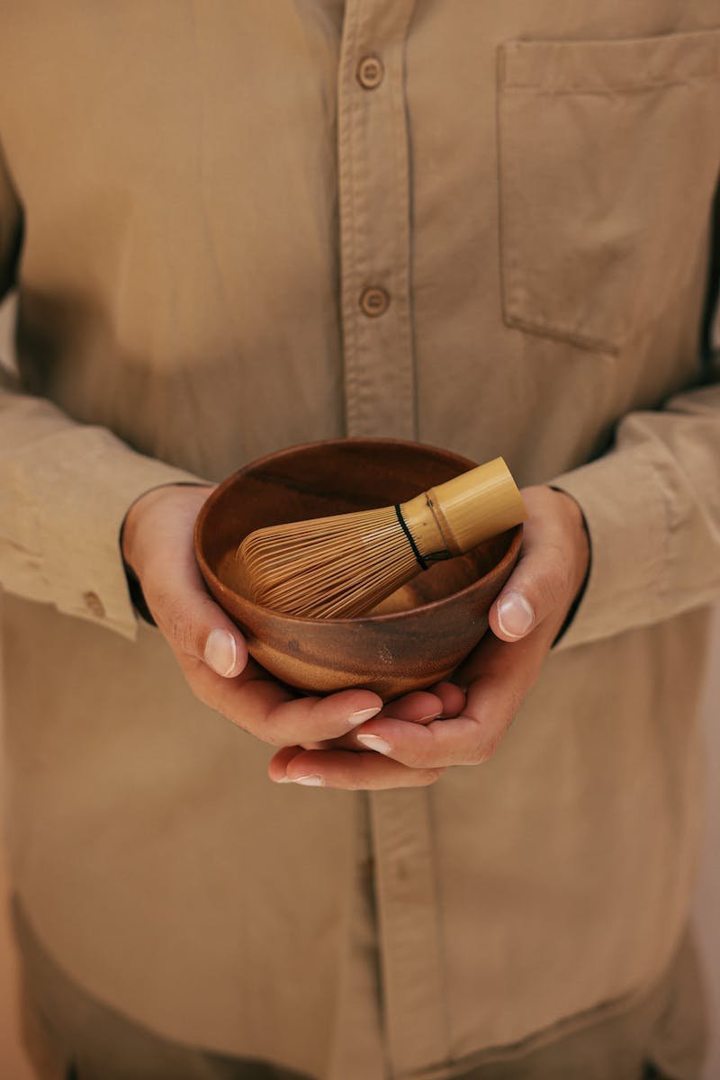 Hands holding bamboo whisk in wooden bowl, ideal for tea ceremony concepts.